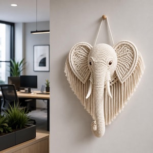 Op de afbeelding: Een crèmekleurige macramé olifantenkop wanddecoratie. De olifant heeft grote, hartvormige oren met ingewikkelde geweven patronen en lange franjes. De slagtanden zijn wit en de ogen bruin. Hangt aan een houten peg.