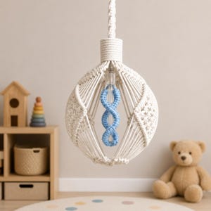 May include: A cream-colored macrame pendant light with a blue, twisted cord center. The light fixture is suspended from a white cord and hangs in a room with a neutral color palette. A wooden shelf with toys and a teddy bear are in the background.