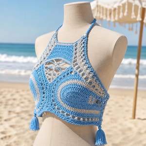 May include: A handmade, blue and beige crochet halter top with an openwork design. The top has a halter neck, tassels, and a detailed pattern. Ideal for beach or summer wear.