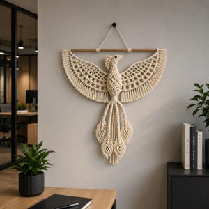 May include: A cream-coloured macrame wall hanging depicting a phoenix with outstretched wings. The bird is suspended from a wooden dowel and features detailed knotting, including a fringed tail and wings. The wall art is displayed against a neutral-coloured wall.
