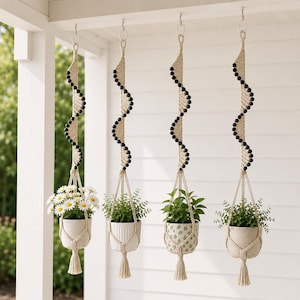 May include: Four hanging macrame plant holders with white ceramic pots. Each holder has a spiral design with black beads and a tassel. The pots contain various green plants and white daisies, creating a decorative display.