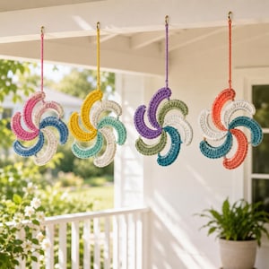 May include: Four colorful, crocheted pinwheels hanging from a white porch ceiling. Each pinwheel features a spiral design with various colors, including pink, blue, yellow, green, and orange. The pinwheels are suspended by matching colored cords.