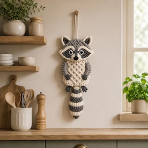 May include: A handmade macrame raccoon wall hanging. The raccoon features a gray and cream color scheme with black accents for the eyes and nose. The tail is striped. The wall hanging is suspended by a cream-colored cord and is a decorative item.
