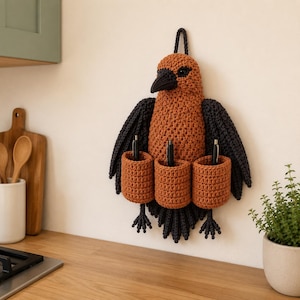 May include: A handmade, crochet bird-shaped wall organizer in shades of brown and black. The bird has three cylindrical pockets for storage and a loop for hanging. Pens are visible in the pockets. The bird is mounted on a light-colored wall.