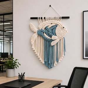 Könnte beinhalten: Makramee-Wandbehang in Form eines Fisches. Der Fisch hat einen cremefarbenen Körper mit blauen Akzenten und eine schwarze Holzstange zum Aufhängen. Der Fisch hat ein detailliertes Design mit geknoteten und gefransten Elementen.