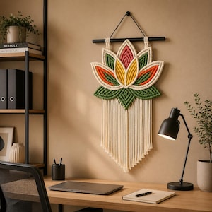 May include: A macramé wall hanging shaped like a lotus flower. The flower has orange, yellow, and pink petals, green leaves, and a cream fringe. It is suspended from a black wooden rod.