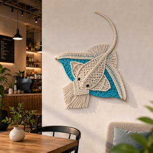 Puede incluir: Un colgante de pared de macramé con forma de manta raya. La manta raya presenta un cuerpo color crema con alas azules y ojos de cuentas negras. La obra de arte mural se exhibe en una pared blanca en un ambiente de cafetería.