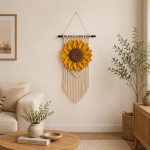 May include: A handmade macrame wall hanging featuring a large, yellow and brown sunflower. The sunflower is the focal point, set against a cream-colored macrame backdrop with fringe. The wall decor is suspended from a black wooden rod.