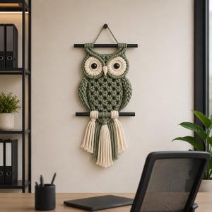 May include: A handmade macrame owl wall hanging. The owl features a green body with cream-colored accents and large black eyes. It is suspended from a black rod and has a fringe of cream and green tassels. The owl is a decorative piece.