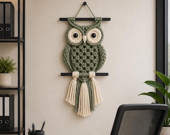 May include: A handmade macrame owl wall hanging. The owl features a green body with cream-colored accents and large black eyes. It is suspended from a black rod and has a fringe of cream and green tassels. The owl is a decorative piece.