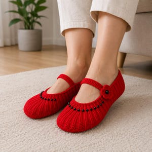 May include: Red crochet Mary Jane slippers with a strap and black bead accents. The slippers have a flower detail on the side and are made of a ribbed knit pattern. These cozy slippers are perfect for indoor wear.