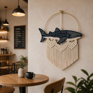Op de afbeelding: Een marineblauwe walvishaai wandhanger met witte stippen, opgehangen aan een gouden ring. De haai bevindt zich boven een crèmekleurig macramé ontwerp met franjes. De achtergrond is een neutrale muur in een café.