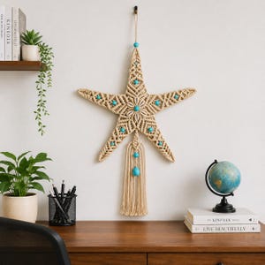 Può includere: Appendiabiti a parete a forma di stella marina in macramè beige con perline turchesi. L'arredamento a tema nautico presenta un design centrale a conchiglia e una nappa. Appeso a una parete bianca sopra una scrivania in legno.