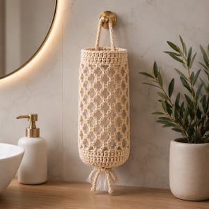May include: A cream-colored, crocheted plastic bag holder with a handle, hanging from a gold-toned hook. The holder has a lattice design and a tied closure at the bottom. It is displayed in a bathroom setting, next to a white soap dispenser and a potted plant.