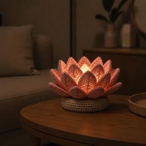 May include: A handmade lotus flower lamp, crafted from pink and cream-coloured yarn. The lamp emits a warm light, highlighting the detailed crochet work. It rests on a circular base and is positioned on a wooden table, creating a cosy atmosphere.