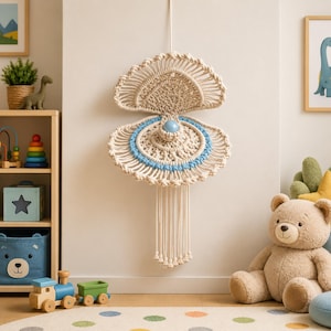 May include: A beige macrame wall hanging with a seashell design. The wall decor features a light blue accent and a central blue bead. The bottom has long, dangling fringe. The wall hanging is displayed in a child's room.