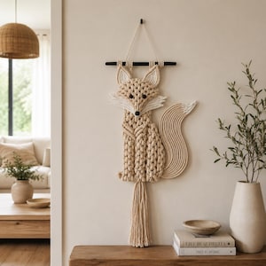 May include: A beige macrame fox wall hanging with a black hanging rod. The fox has a white face and tail tip, with a long tassel at the bottom. The fox is hanging on a beige wall.