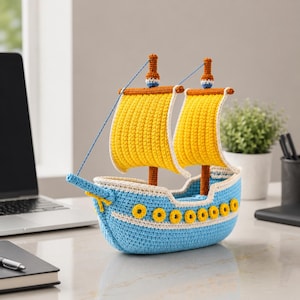 Peut inclure: Un voilier crocheté à la main, bleu, jaune et marron. Le bateau présente des voiles jaunes, une coque bleue avec des détails jaunes et des mâts bruns. La décoration sur le thème nautique est posée sur un bureau.