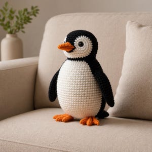 Può includere: Un pinguino all'uncinetto fatto a mano con testa, ali e schiena nere, pancia bianca e becco e zampe arancioni. Il pinguino è seduto su un divano beige. Il pinguino è un giocattolo morbido.