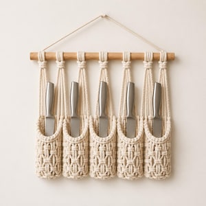 May include: A macrame knife holder with five stainless steel knives. The holder is made of cream-colored woven cotton and hangs from a wooden dowel. Knives are evenly spaced in individual pockets.