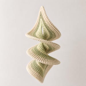 May include: A spiraling, handmade crochet mobile in shades of green and cream. The mobile features a repeating geometric pattern, with each section gradually changing color. The crochet work is detailed, and the overall design is modern and artistic.