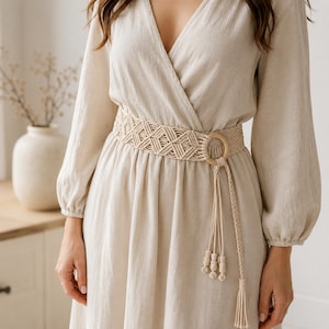 May include: A beige macrame belt with a wooden ring buckle and tassel detail. The belt is worn over a long-sleeved, beige wrap dress. The dress has a V-neck and a flowing skirt. The belt is approximately 3 inches wide.