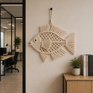 May include: A beige macrame fish wall hanging with a diamond pattern body, fins, and tail. The fish is suspended from a black hook on a beige wall. The fish is approximately 20 inches long.