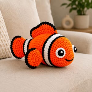 May include: A crocheted orange and white clownfish with black detailing, including fins and eye outlines. The handmade plush toy features large, round eyes and a smiling mouth. It is positioned on a beige sofa.