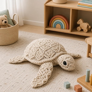 May include: A beige macrame turtle-shaped cushion with a detailed geometric pattern. The turtle has a rounded shell, four flipper-like limbs, and a small head with a black eye. It is placed on a light-coloured rug.