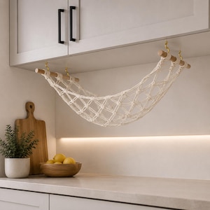 May include: A macrame fruit hammock suspended from a cabinet. The off-white net is held by wooden dowels and gold-colored hooks. A wooden cutting board, a bowl of lemons, and a potted plant are on the countertop below.