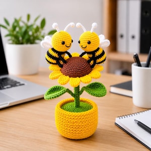 May include: A crocheted sunflower with two smiling bee figures. The flower has yellow petals, a brown center, and green leaves. The bees are yellow and black, with white wings. The flower is in a yellow pot.