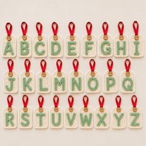Può includere: Un set di etichette alfabetiche all'uncinetto, con lettere dalla A alla Z in verde su sfondo crema. Ogni etichetta ha un anello di nastro rosso e un piccolo bottone di legno. Ideale per regali personalizzati o decorazioni festive.