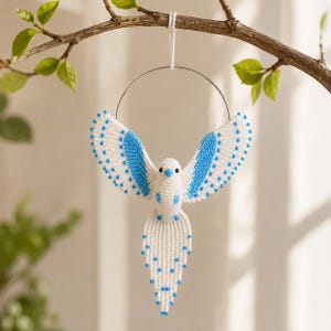 May include: A handmade white and blue macrame bird ornament hanging from a silver metal ring. The bird has outstretched wings with blue accents and blue beaded details. The bird is suspended from a tree branch with green leaves.
