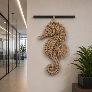 May include: A beige macrame seahorse wall hanging. The seahorse is intricately knotted, with a detailed head, body, and curled tail. It hangs from a black rod, adding a touch of coastal decor to any space. The seahorse is approximately 20 inches tall.