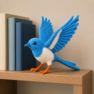 May include: A handcrafted blue and white crochet bird with open wings, displayed on a wooden shelf. The bird has orange feet and a black eye, with books in the background. A decorative item for the home.