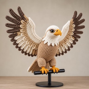 Pode incluir: Uma escultura de águia de crochê com as asas abertas, pousada em um suporte preto. A águia apresenta fios marrons, bege e brancos, com bico e pés laranja. A envergadura é ampla, mostrando um trabalho de crochê detalhado.