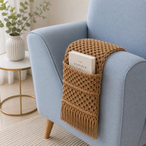 May include: A light blue armchair with a brown macrame armrest organizer. The organizer has a pocket holding a book titled "The Kinfolk Table". The organizer has a fringe at the bottom.