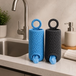 May include: Two handmade crochet bag dispensers. One is bright blue, the other is dark gray. Each has a circular handle at the top and a small opening at the bottom, with a blue bag peeking out. They sit on a white countertop.