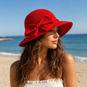 Puede incluir: Un sombrero de sol de ganchillo rojo brillante con un gran lazo. El sombrero tiene un ala ancha y una corona redonda. El sombrero se usa en un día soleado en la playa.
