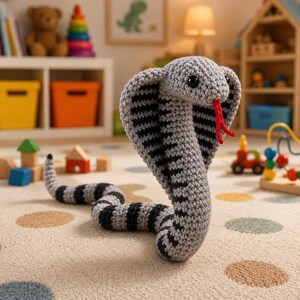 Op de afbeelding: Een gehaakt cobra-slangenspeelgoed in grijs en zwart met een rode tong. De slang staat rechtop, met zijn kap uitgevouwen. Het speelgoed ligt op een patroonkleed in een kinderkamer, met ander speelgoed op de achtergrond.