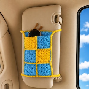 May include: A handmade crochet car organizer in yellow and blue, with a grid pattern. The organizer is attached to a beige car interior panel and holds a pair of sunglasses.