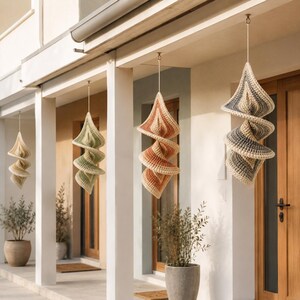 May include: Hanging spiral-shaped crochet decorations in various colors, including beige, green, orange, and gray. The decorative pieces are suspended from a porch ceiling, adding a touch of handmade charm to the outdoor space. The crochet work has a textured appearance.