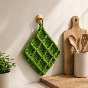 May include: A bright green, diamond-shaped, knitted potholder with a grid pattern hangs from a brass hook on a white wall. A wooden cutting board and utensils in a ceramic holder are in the background, suggesting a kitchen setting.