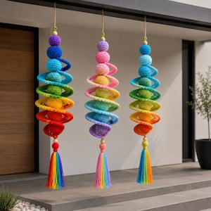 Pode incluir: Três ornamentos em espiral de crochê coloridos, feitos à mão, pendurados em uma viga. Cada ornamento apresenta uma série de esferas e anéis em espiral coloridos do arco-íris, encimados por uma conta de madeira e uma borla. As borlas também são coloridas do arco-íris.