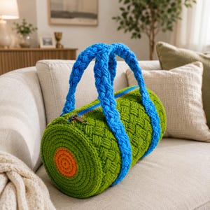 May include: A handmade, green and blue crochet duffel bag. The cylindrical bag features a woven pattern with blue braided handles and a blue zip. The ends of the bag have orange and green concentric circles. The bag is on a cream-coloured sofa.