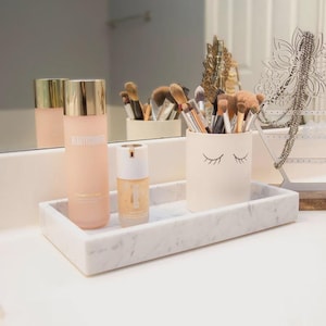 May include: A rectangular white marble tray holds a tall pale pink bottle with a gold cap, a smaller pump bottle, and a cream-colored ceramic cup featuring a black graphic of closed eyelashes, filled with various makeup brushes.