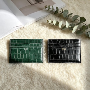 May include: Two rectangular card holders with a crocodile-embossed texture. One is dark green with the gold-colored initials GB, and the other is black with the gold-colored initials DG. Both feature horizontal slots for card storage.