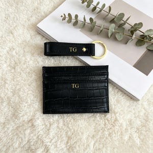 May include: A black card holder and a matching keychain, both featuring a crocodile-embossed texture and the gold-embossed initials TG. The card holder is rectangular, and the keychain includes a gold-toned metal ring and a snap button closure.