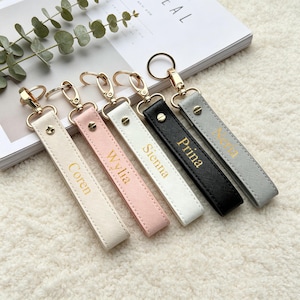 May include: Five rectangular faux leather keychains in cream, light pink, white, black, and gray. Each features a gold-tone metal lobster clasp, a circular ring, and a gold screw stud. The names Coren, Wylia, Sienna, Prina, and Nena are printed in gold.
