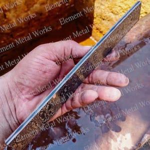 May include: A rectangular metal bar featuring a dark, intricate, etched floral pattern across its surface. The object displays a layered construction visible along its thin side, showcasing alternating light and dark metallic bands in a flat profile.